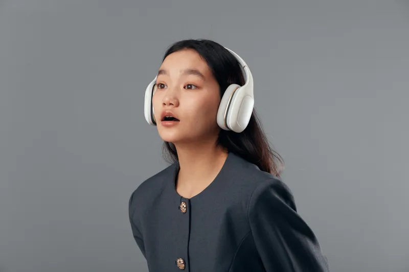 Young woman with long hair wearing headphones and gray jacket, looking amazed or surprised with open mouth on plain gray background. Female listening to music or audio content indoors.