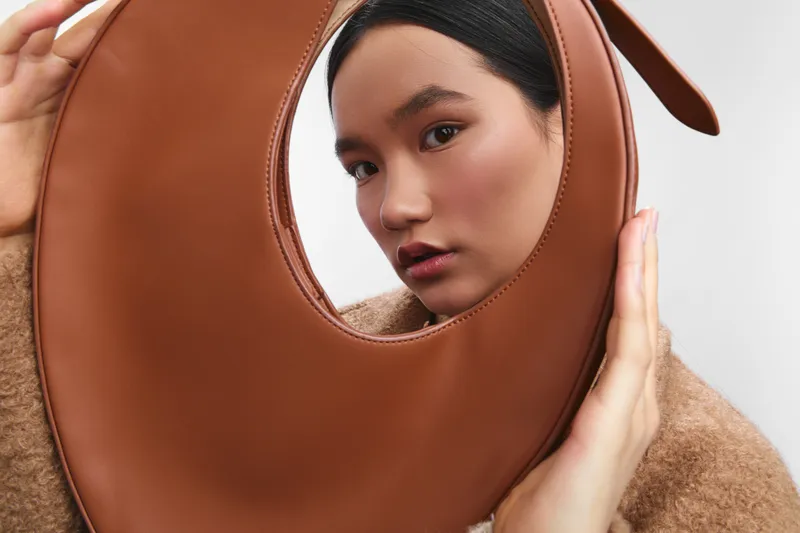 asian woman gazes softly through a brown circular handbag, fashion forward studio shot with clean lighting, elegant expression, contemporary accessory focus, stylish and chic.