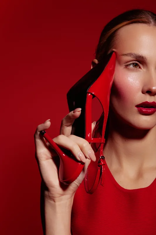 Close-up of a stylish woman holding a red high heel shoe near her face against a red background. Concept of fashion, elegance, beauty, and modern style.