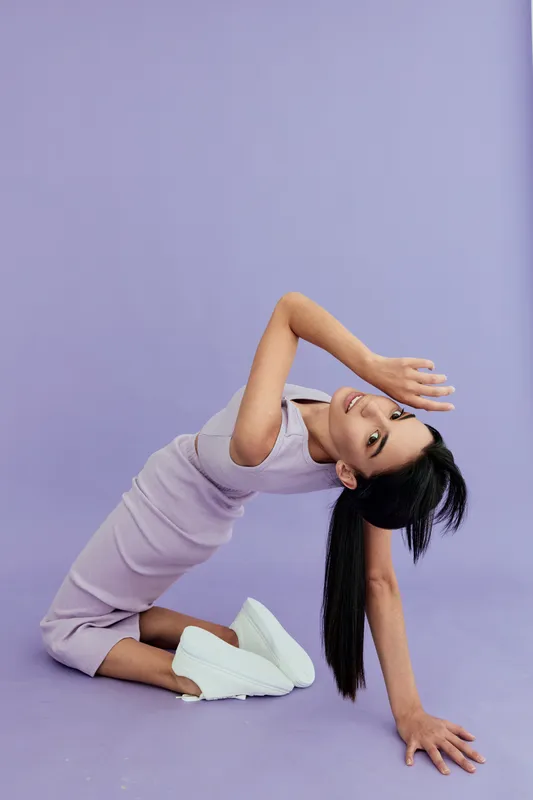 Young woman in lilac dress and white sneakers poses gracefully on purple background, showcasing elegance and modern style with long black hair flowing.