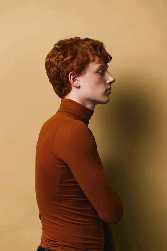 Young redhead man in profile wearing brown turtleneck standing with arms crossed against beige background. Side view portrait of thoughtful male with curly hair indoors.
