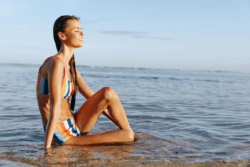 Woman on beach sitting by ocean, relax posture on shoreline, summer sunlight, calm sea waves, fitness mindset, activewear outfit, travel lifestyle portrait, outdoors.