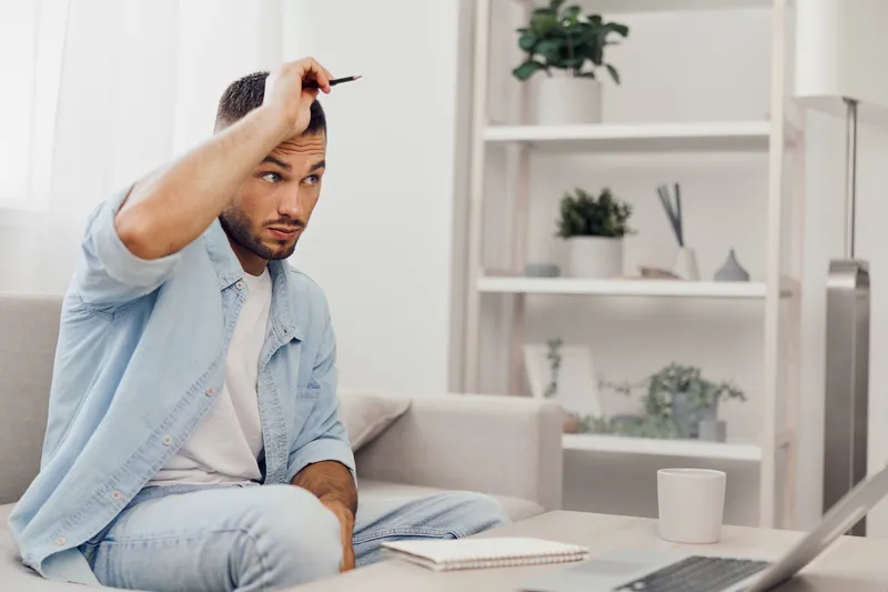 Thoughtful man sitting on a couch with a notebook, brainstorming new ideas while working from home. The contemporary decor adds a modern touch to the scene.