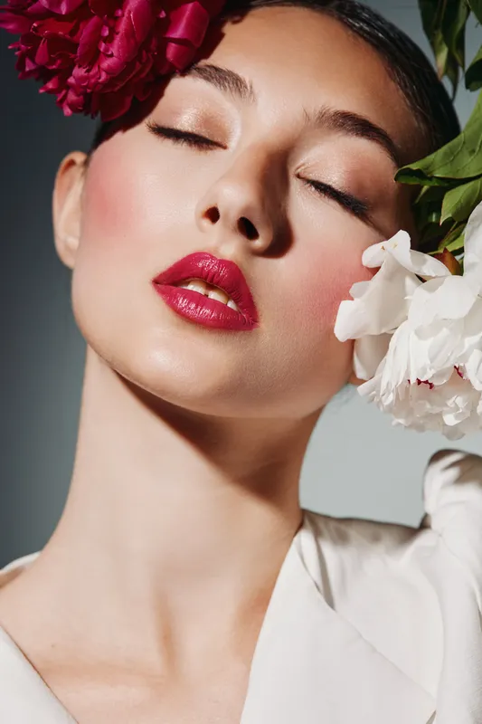 Close-up of young woman with vibrant fuchsia lipstick and flawless skin posing with closed eyes and dramatic pink flowers. Concept of beauty creative fashion, elegance and style.