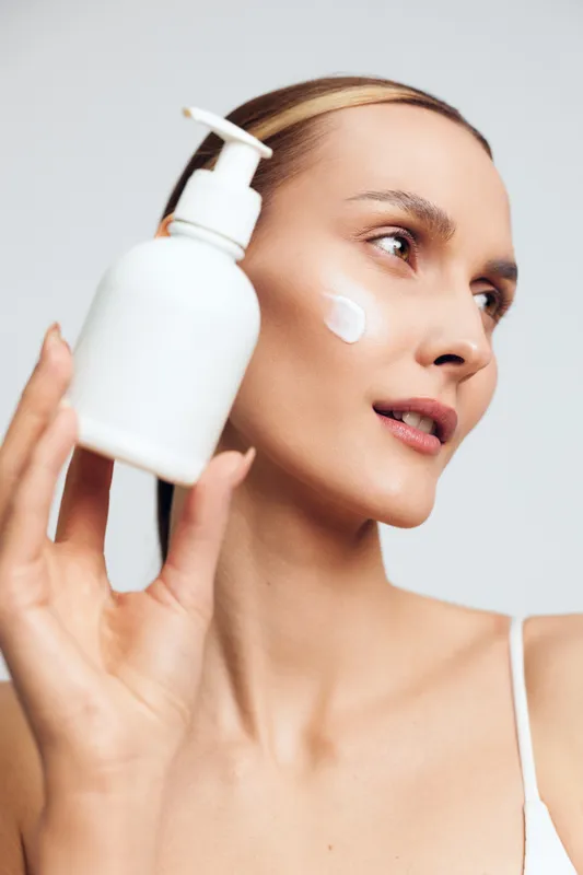 Young woman with clear skin holding a white lotion bottle applying moisturizer on her cheek. Close-up skincare concept with natural light and soft neutral background for beauty and wellness.