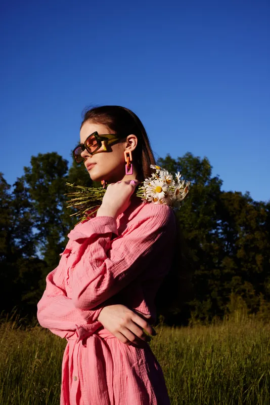 Elegantly dressed young woman in a pink outfit holding wildflowers, exuding confidence and serenity against a bright blue sky and green backdrop