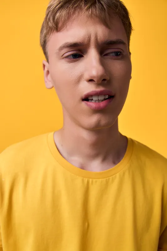 Closeup of a young man in a yellow shirt with a puzzled expression, conveying uncertainty, confusion, and thoughtful mood, studio lighting, vibrant backdrop, casual style, facial tension,