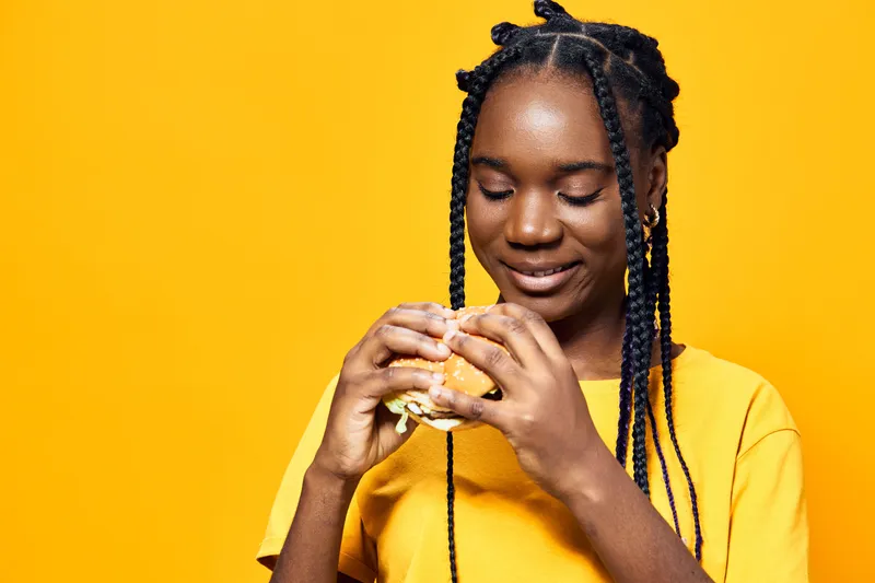 Happy young girl with braids enjoying a delicious burger, wearing a bright yellow shirt against a vibrant yellow background, conveying a joyful eating experience