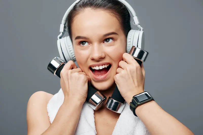Active woman in fitness mode, lifting weights with headphones on, towel around neck, bright studio background, conveying motivation, energy and health through sport.