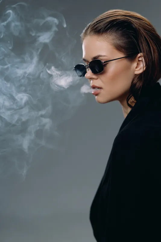 Stylish woman smoking with sunglasses exhaling smoke in dark moody studio lighting.
