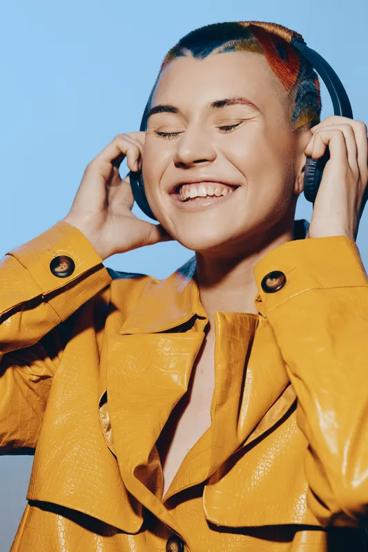 joyful woman with colorful hair enjoys music in large headphones, wearing a bright yellow leather jacket against a clear blue background.