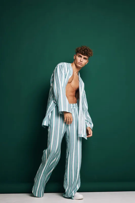 Young man with curly hair wearing striped pajamas standing against green background, casual pose and relaxed expression for fashion or lifestyle concept.