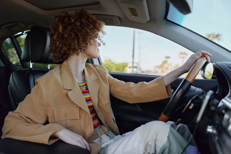 A stylish woman driver sits in the front seat of a car, wearing a beige blazer and striped top. Retro film color gives a nostalgic mood to a casual driving lifestyle.