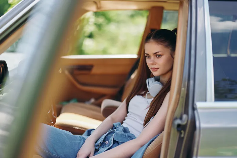 Young woman with long hair relaxing in a car, wearing a white tank top and denim, exuding a calm vibe in a natural green background.