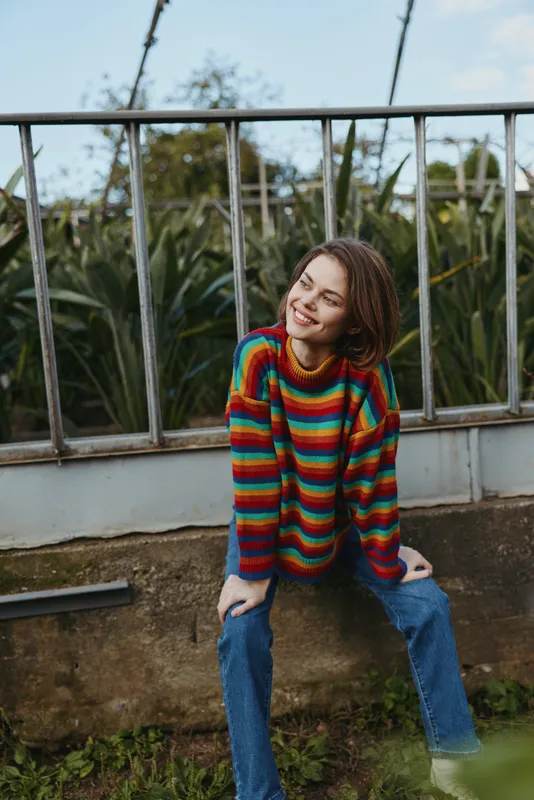 Young woman in a colorful striped sweater smiling while sitting and leaning on a low wall in casual jeans outdoor in an urban garden, relaxed lifestyle portrait and cozy knitwear vibe.