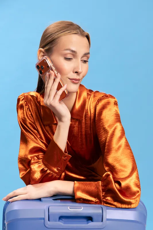 Young woman in shiny orange blouse talks on smartphone while leaning on blue suitcase. Studio portrait on blue background. Concept for travel, communication, and fashion.