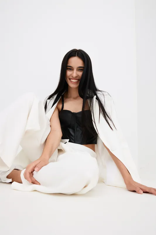smiling woman sitting on white floor in modern white pants and black top with long black hair, natural makeup, bright minimalistic interior, casual fashion style