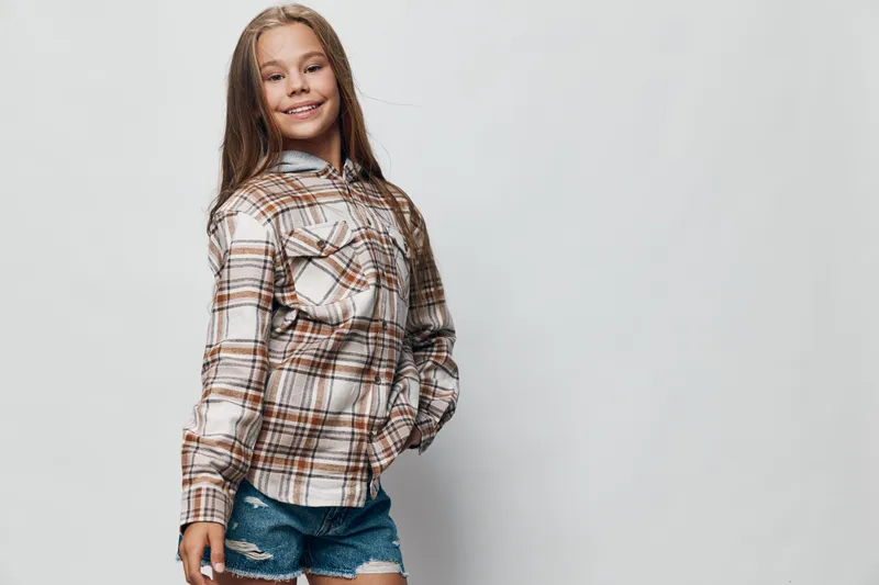 Girl standing with hands in pocket smiling and posing confidently in checkered shirt and denim shorts against light background in studio portrait.