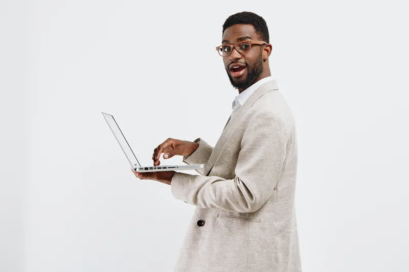 Professional confident young African American man in beige suit holding a laptop and looking at the camera isolated on a plain white background, studio portrait. Business casual style