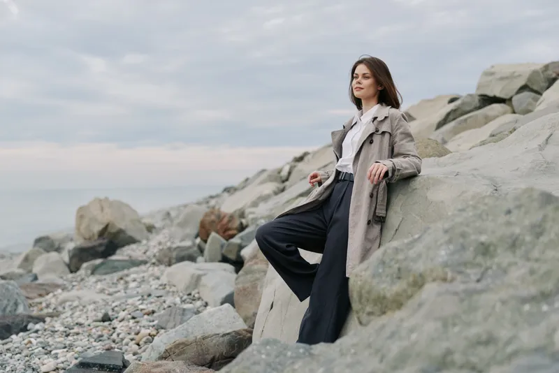Young woman in stylish coat stands on rocky beach, looking away thoughtfully. Overcast sky and natural stones create calm and reflective outdoor atmosphere.