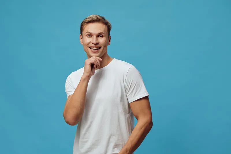 Cheerful smiling young caucasian man in plain white t-shirt looking at camera with hand near chin isolated on vibrant blue background studio portrait. People lifestyle concept.