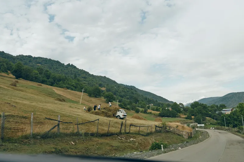 Rural countryside road winding through rolling hills with a weathered fence, a car on a bend, and expansive skies above a quiet scenic landscape.