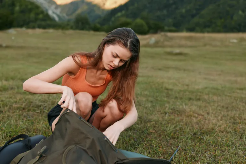 A young woman outdoors is packing a backpack on a grassy field under a clear sky. She wears a sleeveless top and focuses on organizing gear, signaling adventure, preparation, and calm concentration.