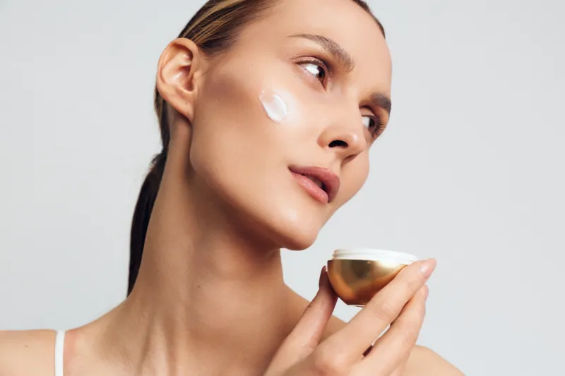 Young woman applying cream on her face holding golden jar. Skincare and beauty concept with clean skin and soft natural light on white background. Healthy lifestyle and self-care.