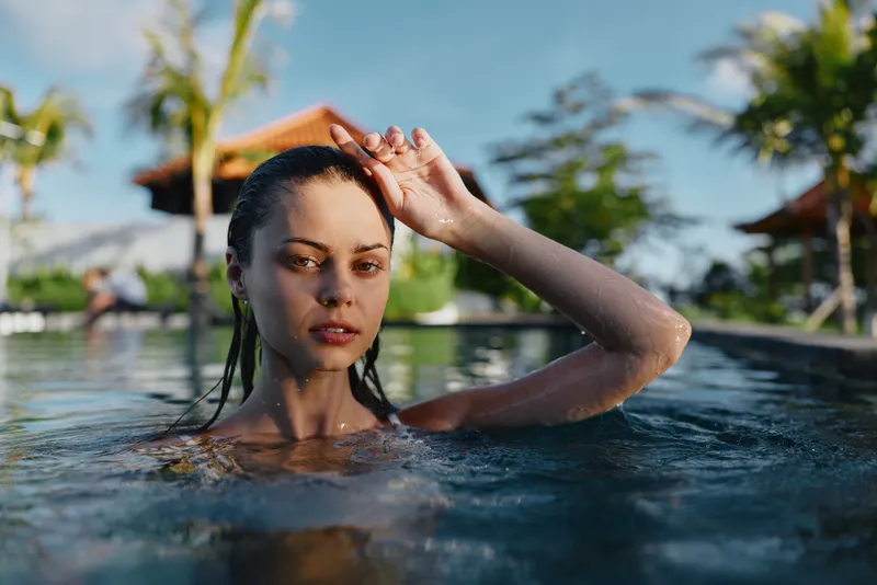 Young woman in a sunlit pool relaxes with hair brushed back, palm trees and umbrellas visible in the background, creating a serene candid moment