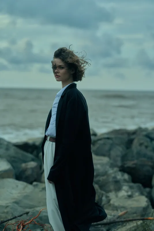 Windy coastal scene featuring a woman in a long coat and light dress standing on rocky shore, contemplating the sea.