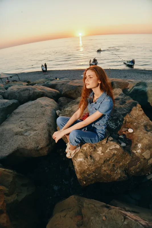 lifestyle woman in modern boho-western denim street style sitting on rocks near ocean during sunset, film color tones enhancing relaxed mood and natural beauty