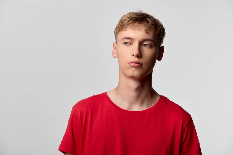 Man, young, in red shirt, looks away in a studio, conveying contemplation and casual style. Minimal backdrop highlights color and relaxed posture, suitable for lifestyle, youth and fashion projects.