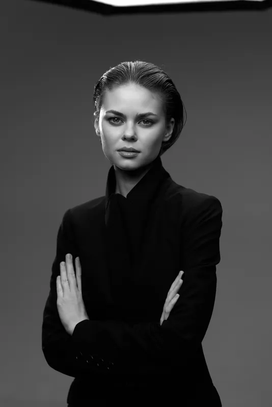 Black and white portrait of a serious woman wearing dark clothing with arms crossed, displaying confidence and strength.