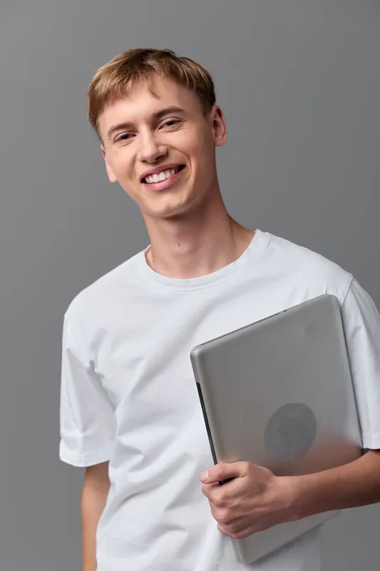 Smiling man holding laptop in a casual studio setting, indoors with gray background, showcasing tech readiness and approachable vibe, perfect for education, business, or startup content