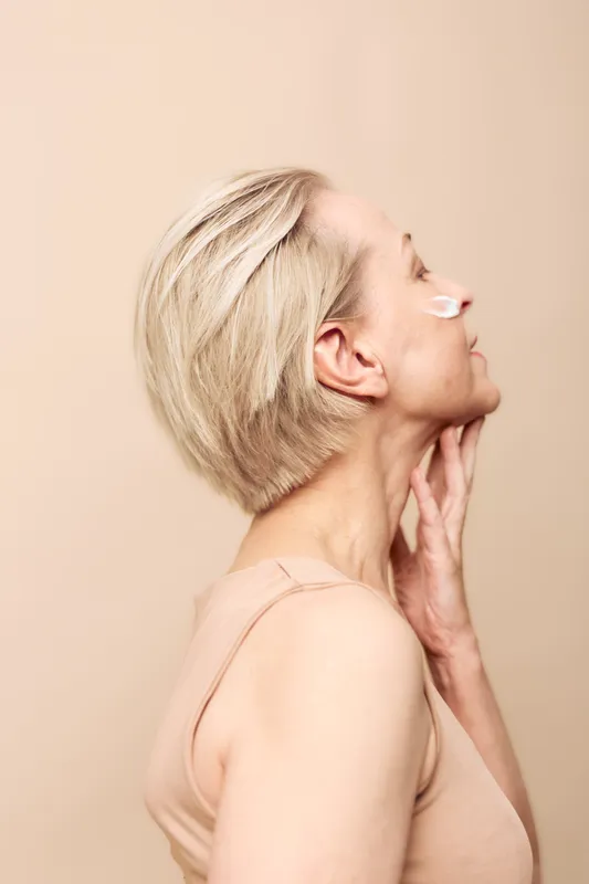 Profile of mature woman with short blonde hair applying cream on face against beige background. Concept for skincare, beauty routine, anti aging, and healthy skin care.