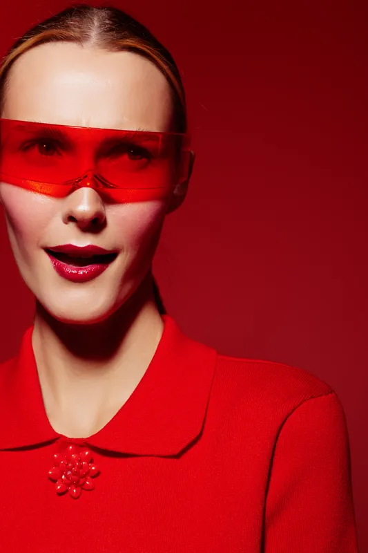 Portrait of a woman wearing red sunglasses and a red top with a collar. The bright red background and glossy lips create a bold, modern fashion concept with strong color contrast.