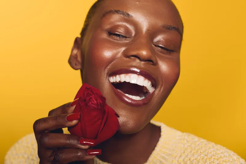 Joyful black woman with dark skin laughing and holding a red rose near her face against yellow background. Concept of happiness, beauty, floral, and natural skincare with bright mood.