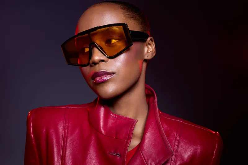 Stylish African American woman with dark skin wearing oversized orange sunglasses and a red leather jacket. Studio portrait with dramatic lighting and confident expression.