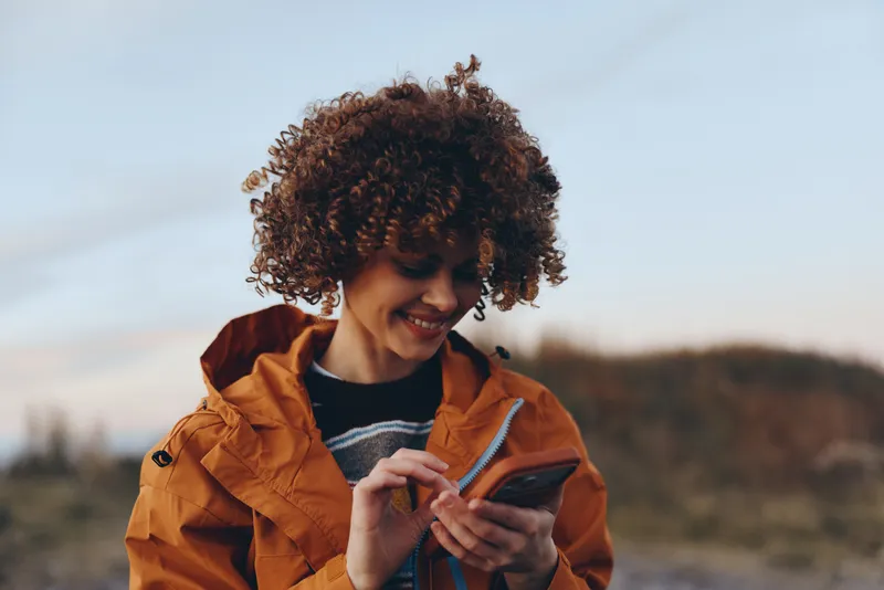 Woman lifestyle outdoors, smiling with curly hair, warm jacket, holding a phone and scrolling messages in gentle sunset light, relaxed candid portrait, blurred landscape, hills