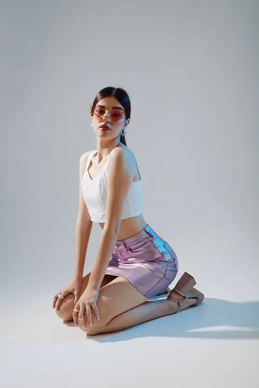 Young woman beauty fashion model kneeling on floor wearing white top and shiny pink skirt with sunglasses. Studio shot with soft lighting on plain background. Concept of style and elegance.