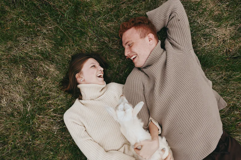Cheerful couple outdoors, lying on grass and laughing with goat kid nearby, cozy sweaters, relaxed mood, sunlit scene, natural colors, candid lifestyle moment, shared affection, outdoors