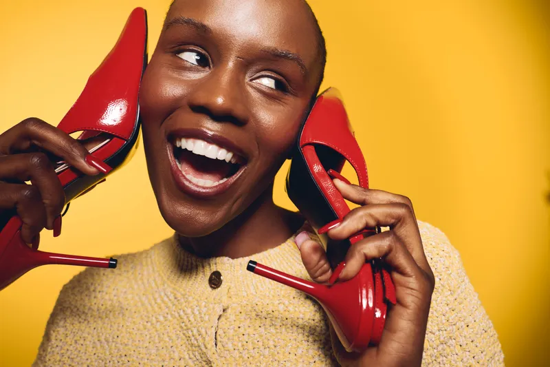 Happy black woman with dark skin holds red high heels near face smiling brightly on yellow background. Concept of fashion, joy, and beauty with African ethnicity and vibrant colors.