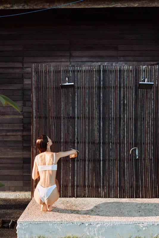 Woman in white bikini kneels under an outdoor shower, relaxing by warm water against a dark wood wall, sunny day, casual poolside mood, serene lifestyle scene with glow.