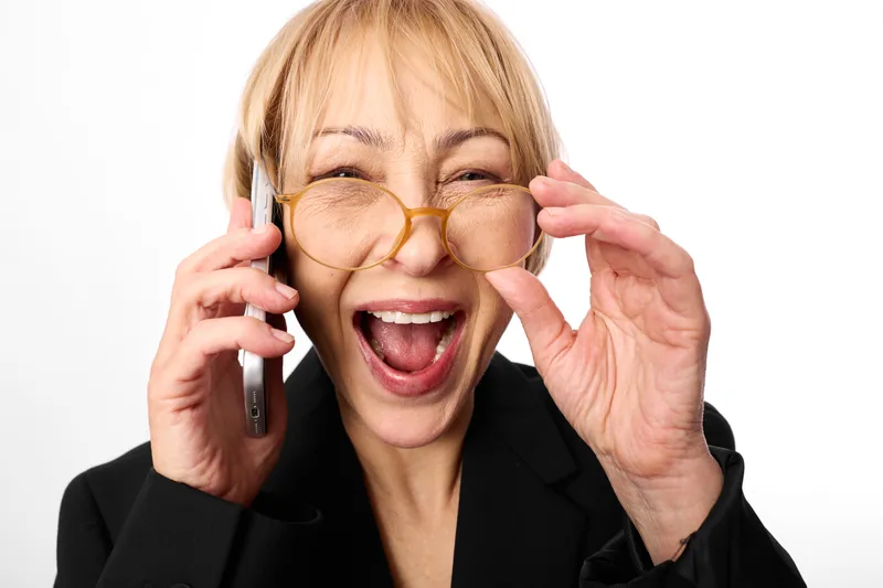 Cheerful woman in business suit, laughing while holding a phone to her ear and adjusting glasses, close up portrait in bright studio light, white background, joyful mood