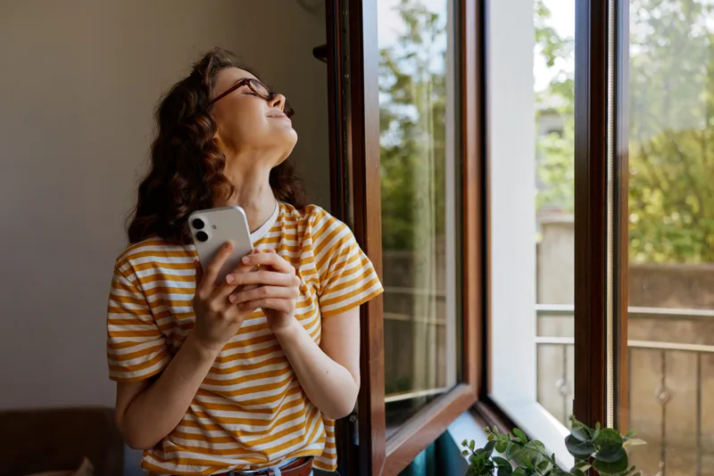A woman in a striped shirt stands by a sunlit window, holding a smartphone and looking up thoughtfully. A candid home lifestyle moment featuring everyday technology use.