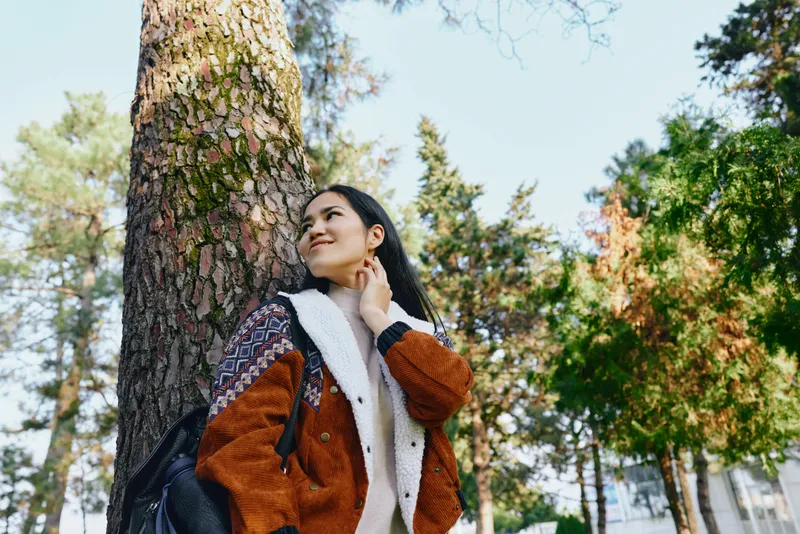 Woman in a warm orange jacket rests against a tall tree in a sunlit park, looking upward with a gentle smile, surrounded by autumn foliage and calm natural scenery