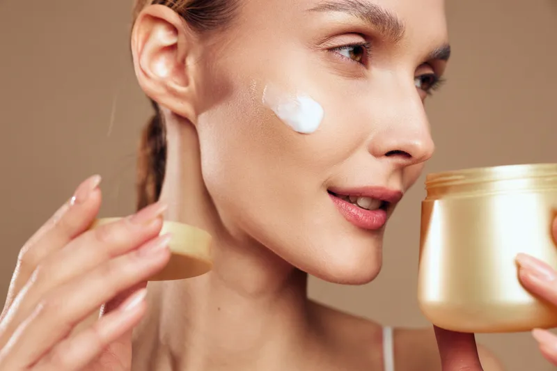 A young woman applies moisturizing cream on her cheek while holding an open jar. The close-up shows smooth skin and a neutral background, concept for skincare and beauty routine.