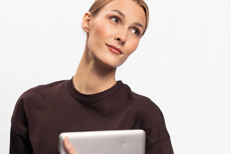 Young woman in brown sweater holding tablet looks thoughtful with slight smile on white background. Concept of technology, casual lifestyle, and modern communication.