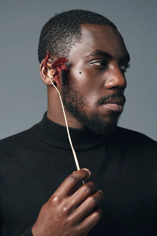 African man with dark skin holding dried flower near face in creative conceptual visual style. Portrait of thoughtful black male model wearing black turtleneck on gray background.