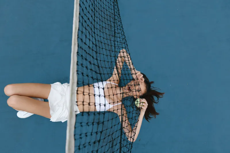 Young athletic woman in sporty attire expressing relaxation under a tennis net, showcasing a calm summer vibe with a vibrant blue background